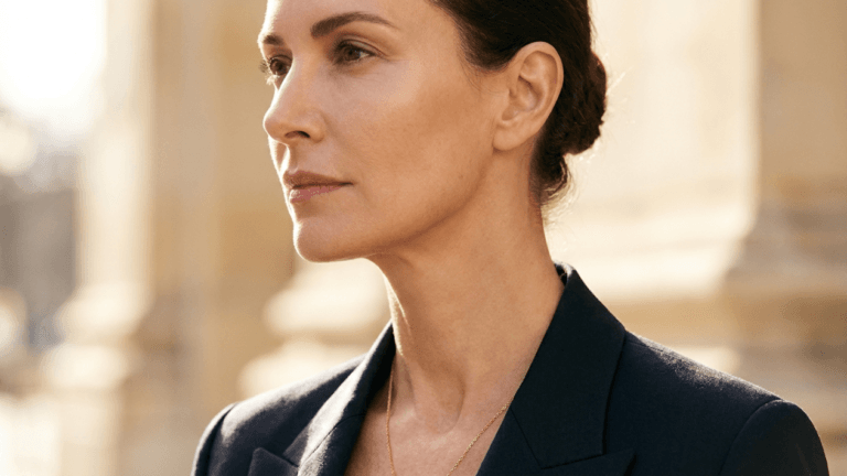 A professional, high-end editorial portrait of a woman in her prime, captured with an 85mm prime lens for a shallow depth of field, rendering the image in hyper-realistic 8k resolution. The lighting is cinematic, utilizing soft, natural golden-hour backlighting that creates a delicate rim light effect along her jawline and profile, highlighting her refined bone structure and elegant, poised posture. Her skin texture is portrayed with exquisite natural clarity, featuring a healthy, radiant luminescence without heavy retouching. She is dressed in a sharp, tailored dark navy blazer made of high-quality wool-blend fabric, complemented by a minimalist, thin gold chain necklace that catches the light. The composition focuses on her confident, contemplative profile, set against a softly blurred, warm-toned architectural background that evokes a sophisticated, luxurious, and timeless urban atmosphere.