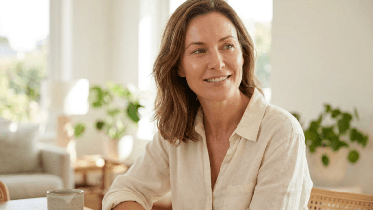 This high-resolution, 8k hyper-realistic portrait is captured with an 85mm lens, showcasing a shallow depth of field that keeps the focus sharply on a poised woman in her mid-forties. The lighting is soft, diffused, and natural, emanating from the side to gently contour her facial features and highlight her radiant, smooth complexion with a healthy, subtle glow. She sits in a relaxed, serene posture, wearing a casual, textured beige linen shirt that drapes elegantly. Her expression is warm and contemplative, gazing slightly off-camera. The background features a bright, airy, and minimalist interior setting, with a hint of lush greenery in a ceramic pot, creating an atmosphere of sophisticated, tranquil, and modern domestic comfort.