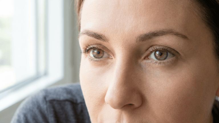 A hyper-realistic, close-up portrait of a woman's face, captured with a professional 85mm macro lens to emphasize intricate skin texture and fine details. The lighting is soft and natural, emanating from the side as if from a window, creating gentle highlights on the cheekbones and subtle shadows that add depth to her features. The subject, a woman with expressive brown eyes, has a serene and contemplative gaze directed slightly off-camera. Her skin appears naturally hydrated, with visible pores and realistic micro-textures that attest to 8k resolution quality. She is wearing a simple, dark-colored textured top. The background is a soft, out-of-focus interior, likely near a window, creating a serene, luminous, and clean atmosphere that highlights the natural beauty and clinical precision of the portrait.