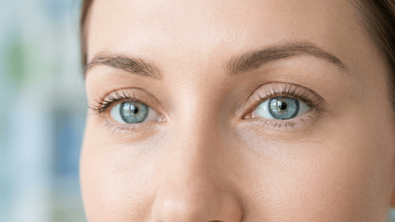 An ultra-high-definition, hyper-realistic macro close-up of a woman's eyes, captured with an 85mm prime lens to achieve clinical precision and shallow depth of field. The lighting is soft and diffused, emphasizing the natural luminescence of the skin and the intricate, cerulean-grey iris patterns. The subject displays a calm, focused gaze, with visible micro-texture of the epidermis, refined pores, and natural lash definition. The composition centers on the eyes, with a neutral, out-of-focus background that enhances the subject's features. The overall atmosphere is clean, medical-grade, and sophisticated, reflecting high-end cosmetic or dermatological editorial standards. No clothing or hardware is visible in this frame, focusing entirely on facial aesthetics and ocular clarity.