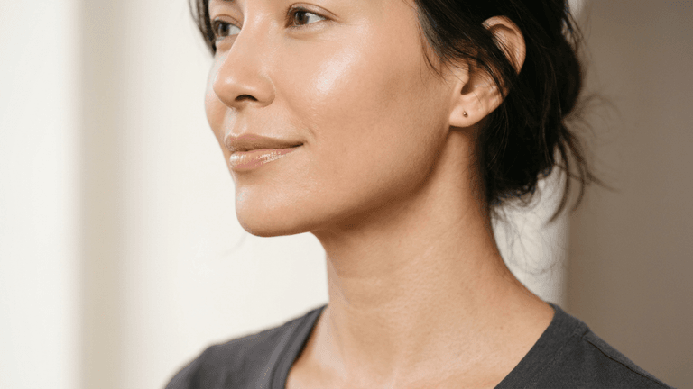 A hyper-realistic 8K editorial close-up portrait of a woman captured with an 85mm macro lens, showcasing a shallow depth of field. The subject is positioned in a serene three-quarter profile, highlighting refined facial architecture and a smooth, elegant jawline. The lighting is soft and natural, emanating from the side to create subtle, flattering contours across the cheekbone and neck without harsh shadows. Her skin texture is impeccable, rendered with a luminous, hydrated glow that captures fine highlights on the bridge of the nose and the cupid's bow. She wears a minimalist charcoal-grey cotton garment, keeping the focus entirely on the sophisticated, clean aesthetic of her complexion and relaxed, poised demeanor against a muted, soft-focus background.