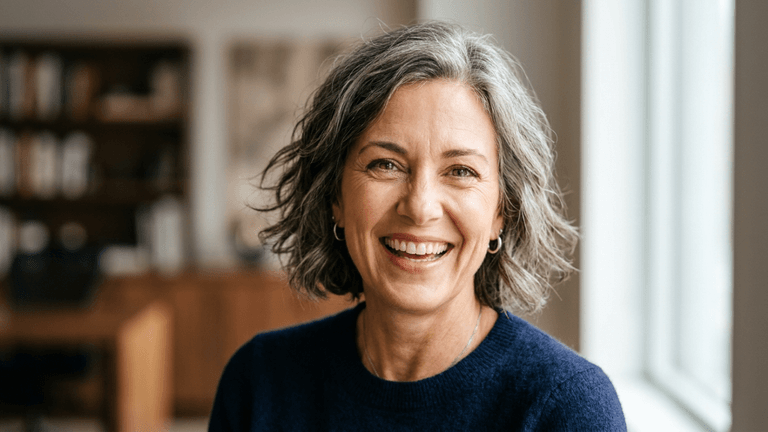 A high-resolution, hyper-realistic portrait of a mature, elegant woman with radiant, silver-toned hair, captured with an 85mm portrait lens to achieve a shallow depth of field. The lighting is soft and natural, streaming from a side window, which gently highlights her expressive facial features and the refined texture of her skin, emphasizing her genuine, joyful expression. She is wearing a dark blue, finely textured knit sweater that adds a sophisticated, understated contrast to the bright background. The composition is a professional head-and-shoulders shot, set against a softly blurred, warm-toned office interior, evoking a professional yet inviting atmosphere. The image quality is exceptional, rendering fine details in her hair, skin luminescence, and the subtle glint of her small silver hoop earrings with crystalline clarity.